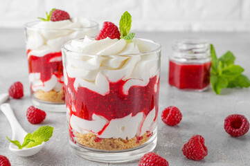 Raspberry trifle with whipped cream, berry sauce, cookie crumbs and mint leaves in a glass on a gray concrete background. Summer diet dessert.