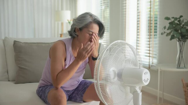 Asia adult people middle age old woman sit at sofa home feel hot flash fever use fan waving hand blowing wind face elder health care summer heat aging issue. Sad pain sweat tired stroke suffer stress.