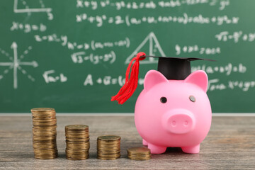 Piggy bank with graduation hat and coins on table near green blackboard. Student loan concept