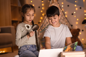 Cute children with magnifier reading adventure book at home in evening