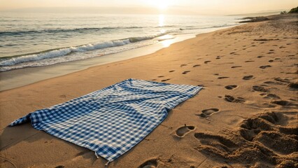 Blue checkered picnic blanket on an empty beach with footprints in the sand during a stunning sunrise