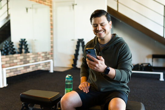 Latin man resting on bench, checking smartphone following workout at gym