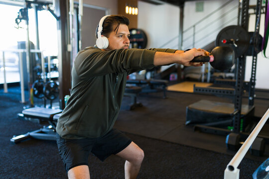 Mid adult man swinging kettlebell while wearing headphones during intense strength training in contemporary gym