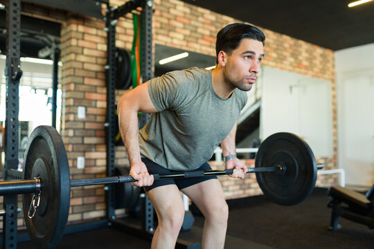 Determined hispanic man lifting a barbell while performing bent-over rows in a modern gym, showcasing strength and dedication to fitness