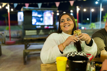 Smiling plus size woman enjoying tasty street food burrito near illuminated food truck, capturing casual urban dining atmosphere after dark