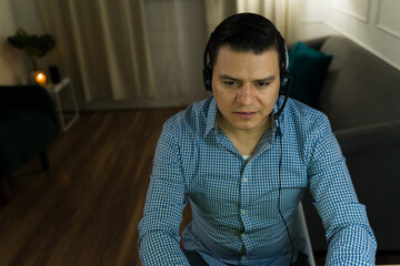 Fototapeta premium Latin man wearing headset working remotely from home at night, focused on his job as sales representative or customer service agent