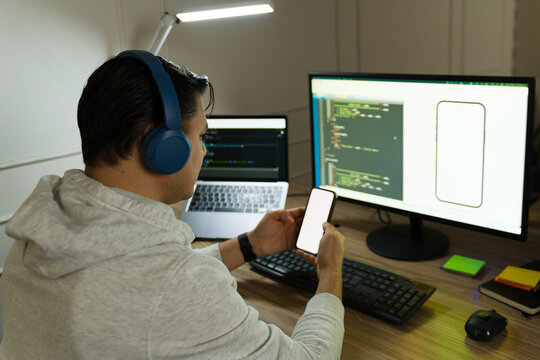 Male programmer wearing headphones working at home comparing smartphone app layout with code displayed on computer screen