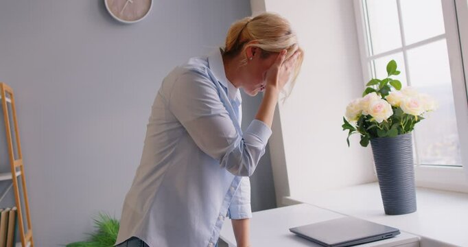 Tired blonde woman standing near window, touching forehead with hand, feeling bad, suffering from dizziness. Sick girl having vertigo, feeling exhausted, overheated or unwell, losing consciousness.