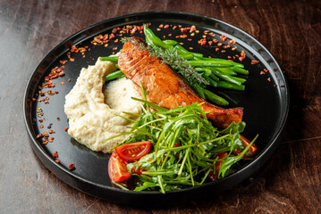 A beautifully plated dish featuring grilled salmon, sautéed green beans, creamy mashed potatoes, and fresh salad with cherry tomatoes, garnished with herbs and spices.
