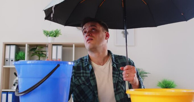 Man at home holding umbrella, having hole or crack in ceiling through which liquid flowing out of roof, in need of professional roofer, sad with necessary office room repairs to find and fix leaks