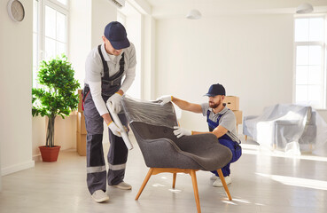 Two professional movers in uniform carefully unpacking and setting up armchair in bright spacious...