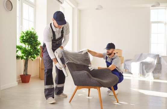 Two professional movers in uniform carefully unpacking and setting up armchair in bright spacious room. Loaders providing relocation service, handling furniture and organizing home moving process.