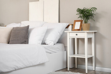 Bedside table with photo frame and houseplant near white wall in interior of bedroom