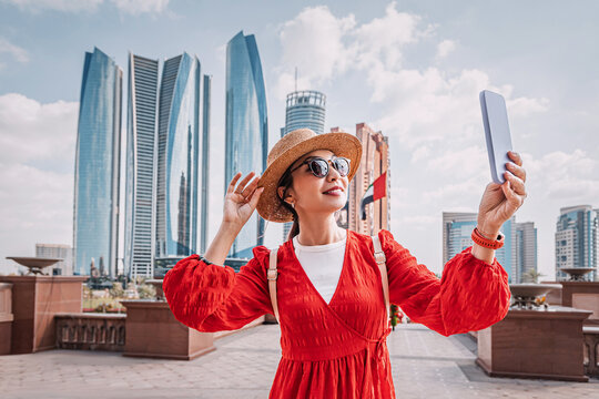 Asian woman wearing red dress and straw hat taking a selfie with smartphone in front of skyscrapers in Abu Dhabi - Powered by Adobe