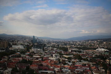 Georgia, A panoramic view of a vibrant city blending historical architecture with modern skyscrapers, set against a backdrop of rolling hills and mountains.