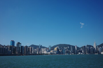 Fototapeta premium A stunning city skyline with towering skyscrapers set against lush mountains, viewed from across the water. The deep blue sky and shimmering waterfront highlight the modern architecture.
