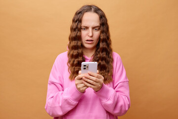 A woman featuring long, wavy hair stands against a brown backdrop. She wears a pink hoodie and...