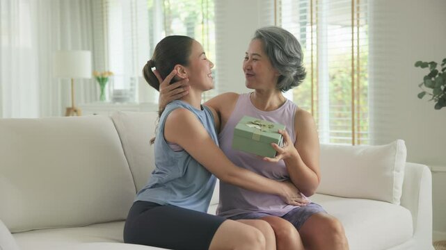 May Mother's day young adult child arm around cuddle hug give gift box to mature middle age old mum. Love kiss care mom asia elder people sit at home sofa happy smile enjoy family good time aging life