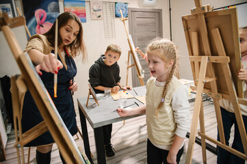 A teacher explains to the children how to draw picture in art class. A group of school children with brushes and an easel in art lesson began drawing landscapes. Children enjoy painting in a workshop.