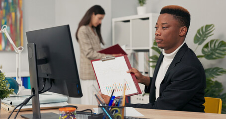 dark-skinned office worker sits in front of computer at desk having video conversation internet connection via webcam discussing company matters with co-workers explaining completed chart project