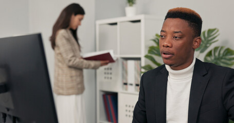Fototapeta premium dark-skinned office worker sits in front of computer at desk having video conversation internet connection via webcam discussing company matters with co-workers explaining completed chart project