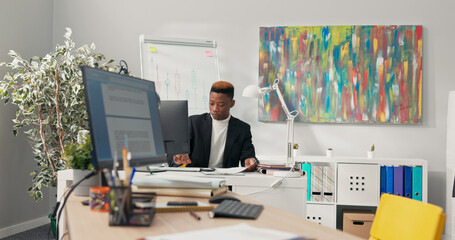 The manager, boss, office director with dark skin dressed in black suit sits behind desk prepares documents for mailing asks assistant to pass envelopes girl fulfills request and returns to her duties