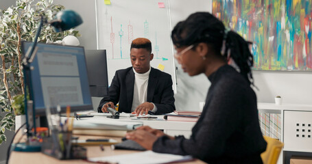 Dark-skinned boss sits on chair with feet on desk, holding tablet in hands, surfing the Internet, puts down equipment, put hands behind head, takes break relaxes closes eyes secretary is still working
