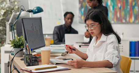 Obraz premium An attractive girl wearing glasses works at a desk in front of a computer, fills out documents, signs checks, in the background co-workers are browsing the project on the computer, talking, discussing