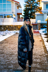 Obraz premium Woman walking down snowy path in winter. A woman dressed in a black puffer coat and hat walks along a snowy path lined with houses on a sunny winter day.