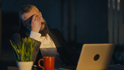 Businessman in a suit working late at night, showing exhaustion and deep thought in a dimly lit...