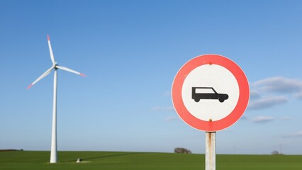 A road sign prohibiting vans is seen in the foreground, with a wind turbine in the background against a clear blue sky. The image combines elements of transportation and renewable energy
