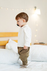 Fototapeta premium Child plays on bed with toy mustache. A young boy stands on a bed, wearing a white shirt and green pants, playing with a toy mustache in a cozy room.
