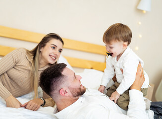 Fototapeta premium Family bonding on a cozy bed. A joyful family moment features a playful child sitting on a dad's lap, while mom smiles nearby.