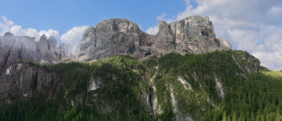 Drone panorama of the Dolomites near Colfosco, Italy, showcasing dramatic rock formations, lush pine forests, and the source of the Pisciadu waterfall