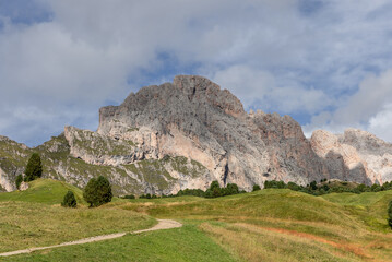 A winding path cuts through green meadows toward the imposing Seceda ridge in South Tyrol, Italy. The towering rocky cliffs and scattered trees create a tranquil alpine atmosphere