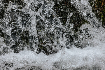 A detailed close-up of foamy bubbles forming on the surface of a fast-moving stream. The intricate texture and reflections capture the dynamic motion of water in nature.