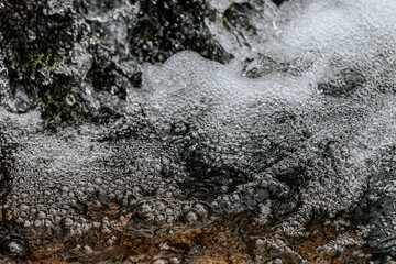 A detailed close-up of foamy bubbles forming on the surface of a fast-moving stream. The intricate texture and reflections capture the dynamic motion of water in nature.