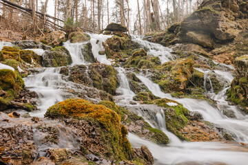 A beautiful multi-tiered waterfall flows over moss-covered rocks in a forest setting. The long exposure captures the smooth movement of the water, creating a peaceful and serene natural scene.