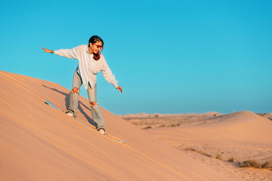 Young woman sandboarding down a sandy dune in the desert, enjoying the thrill of the sport during a stunning sunset