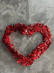 A red heart-shaped wreath made of shiny decorations against a textured gray background.