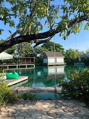 A tranquil waterside scene featuring a small white house, lush greenery, and calm waters reflecting the blue sky.