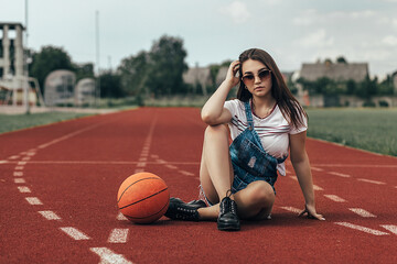 A girl sits on a treadmill with a basketball. She is wearing a white T-shirt with stripes and denim...
