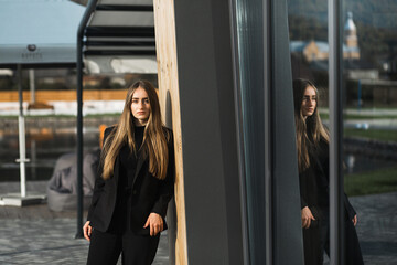 A woman with long straight hair is leaning against the wall. She is wearing a black suit with a jacket and trousers, as well as black high-heeled shoes.