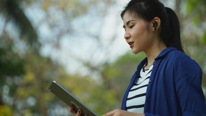 Modern Asian woman engaged in browsing her tablet, embodying a casual yet professional lifestyle,...