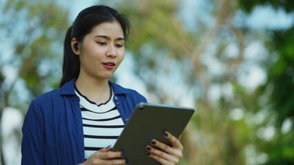 Fototapeta premium Asian Freelancer enjoying the outdoors while connecting digitally through her device, illustrating the future of work and the relaxed lifestyle of modern professionals in a city park.