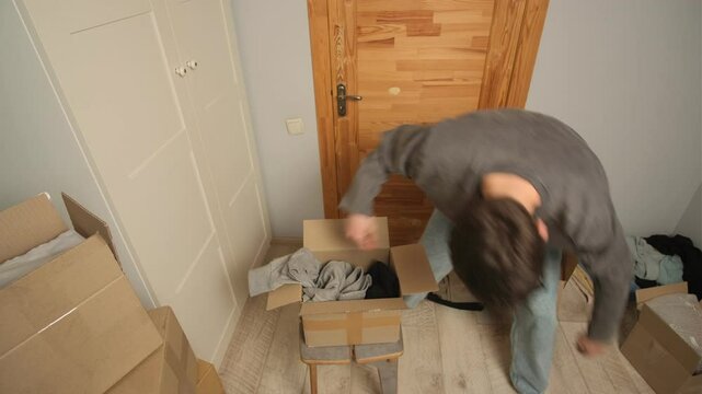 Man in a living room hurriedly and nervously packs clothes into a cardboard box, tossing items with frustration. The space is filled with various boxes, suggesting an unplanned eviction or forced move