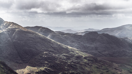 Breathtaking view of expansive mountain ranges shrouded in atmospheric clouds, showcasing rugged terrain and contrasting light at midday. The remote wilderness invites exploration.