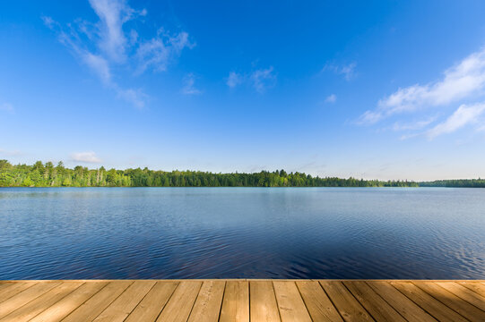 A summer day with a calm blue lake and a wooden dock in the foreground in Algonquin Provincial Park, Ontario, Canada. The water reflects the clear sky, surrounded by lush green trees along the shore. - Powered by Adobe