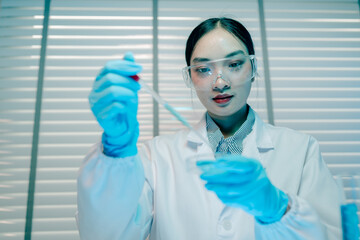 Woman in a lab coat is holding a beaker of blue liquid. She is wearing gloves and a lab coat