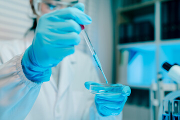 Woman in a lab coat is holding a beaker of blue liquid. She is wearing gloves and a lab coat